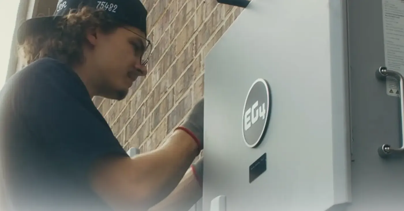 An installer wiring an EG4 hybrid inverter and battery cabinet on the side of a house